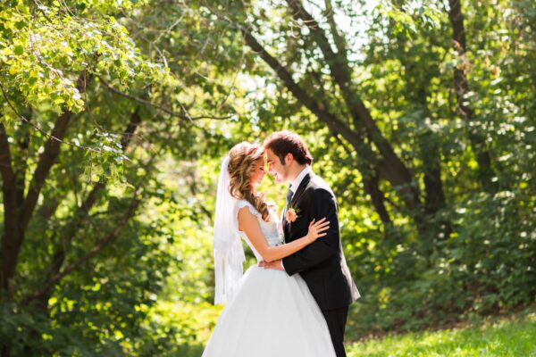Beautiful wedding couple in park. They kiss and hug each other A bride and groom touching foreheads in a bright, grassy meadow in front of a green wooded area.
