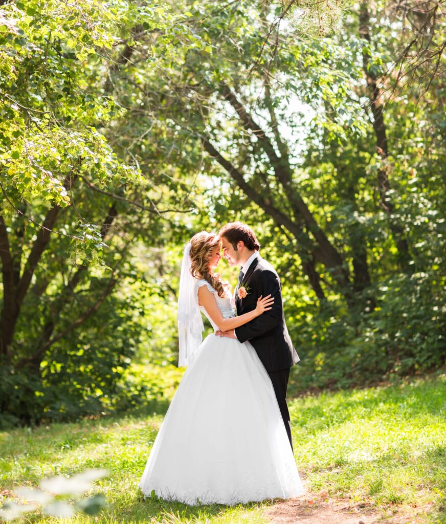 A bride and groom touching foreheads in a bright, grassy meadow in front of a green wooded area.