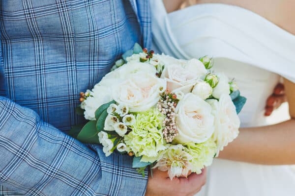 AdobeStock_1223563742smol A bride and groom embracing with a bouquet of flowers between them.