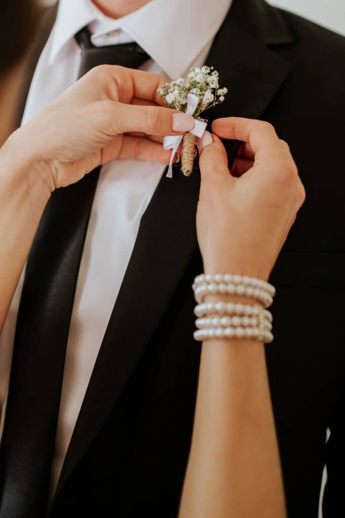 A close-up shot of a bride’s hands pinning a delicate boutonniere onto the groom’s black suit. The floral detail adds an elegant touch, symbolizing love and tradition in wedding ceremonies.