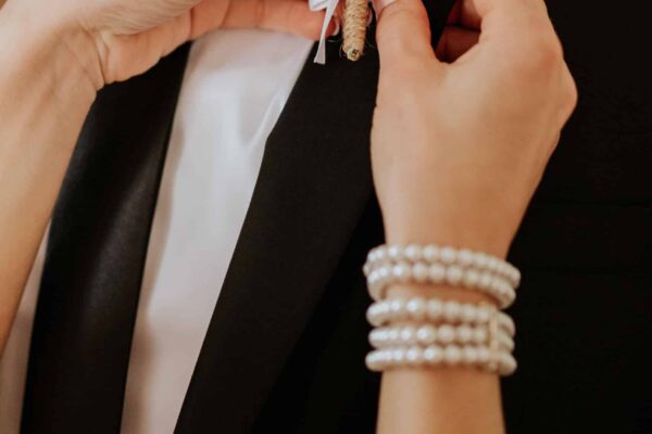 Bride Attaching Boutonniere to Groom’s Suit A close-up shot of a bride’s hands pinning a delicate boutonniere onto the groom’s black suit. The floral detail adds an elegant touch, symbolizing love and tradition in wedding ceremonies.