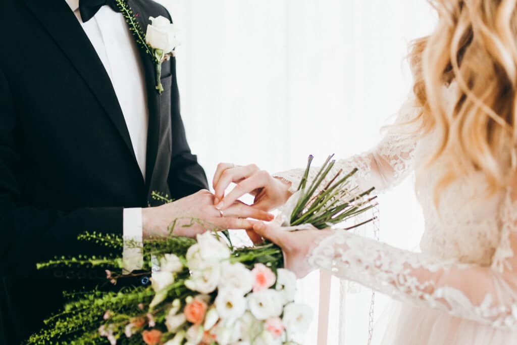 A bride in white slipping a wedding band onto the groom's finger.