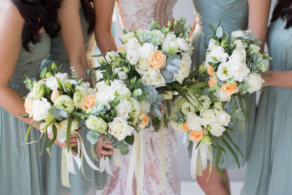 Bridesmaids in blue-grey dresses and a bride in white all holding white, pink and yellow bouquets.