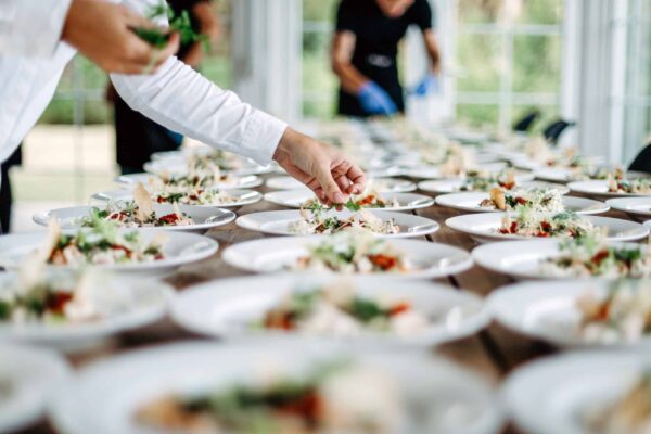 AdobeStock_243451238 A table filled with plates of food being prepped and garnished by event staff.