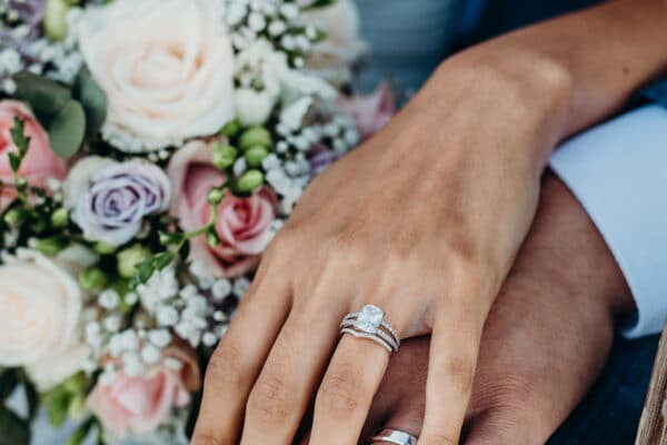 AdobeStock_243451985_web Hands of a bride and groom on top of one another, showing off their wedding rings, with the multicolored bouquet in the background.