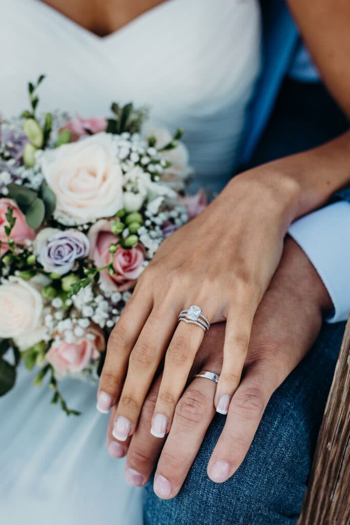 Hands of a bride and groom on top of one another, showing off their wedding rings, with the multicolored bouquet in the background.