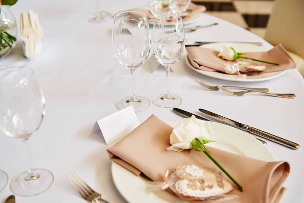 Table setting with blank guest card, white plate with beige serviette, rose, heart and cutlery on table, copy space. Place setting at wedding reception. Table served for wedding banquet in restaurant