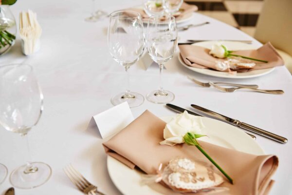 Table setting with blank guest card, white plate with beige serv Table setting with blank guest card, white plate with beige serviette, rose, heart and cutlery on table, copy space. Place setting at wedding reception. Table served for wedding banquet in restaurant