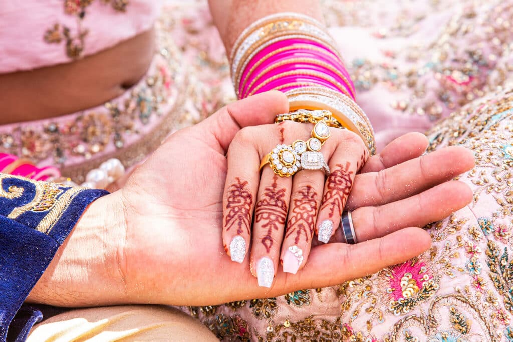 A woman in traditional Indian vestments, including a vibrantly decorated sari, bold jewelry, and henna tattoos, placing her hand in the groom's hand.