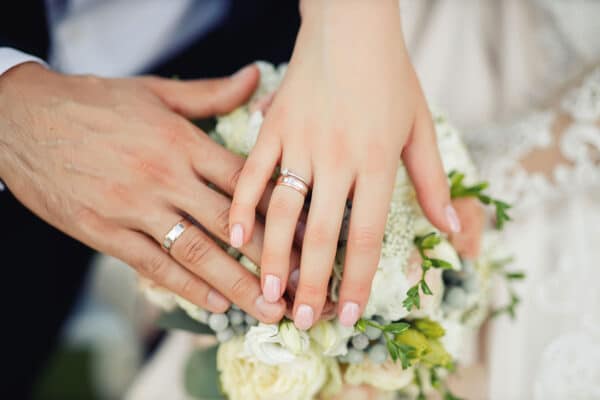 AdobeStock_424677148_web A bride and groom holding their hands out over a white and yellow bouquet, showing off their wedding rings.