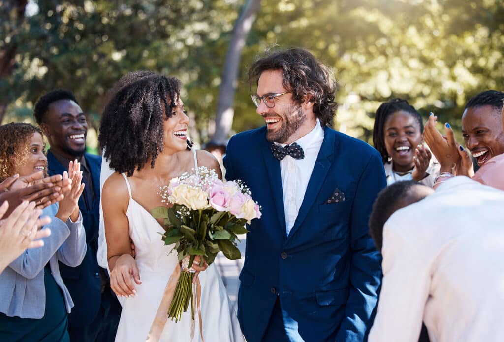 Bride and groom looking at each other and smiling as they leave their wedding, with friends and family cheering.