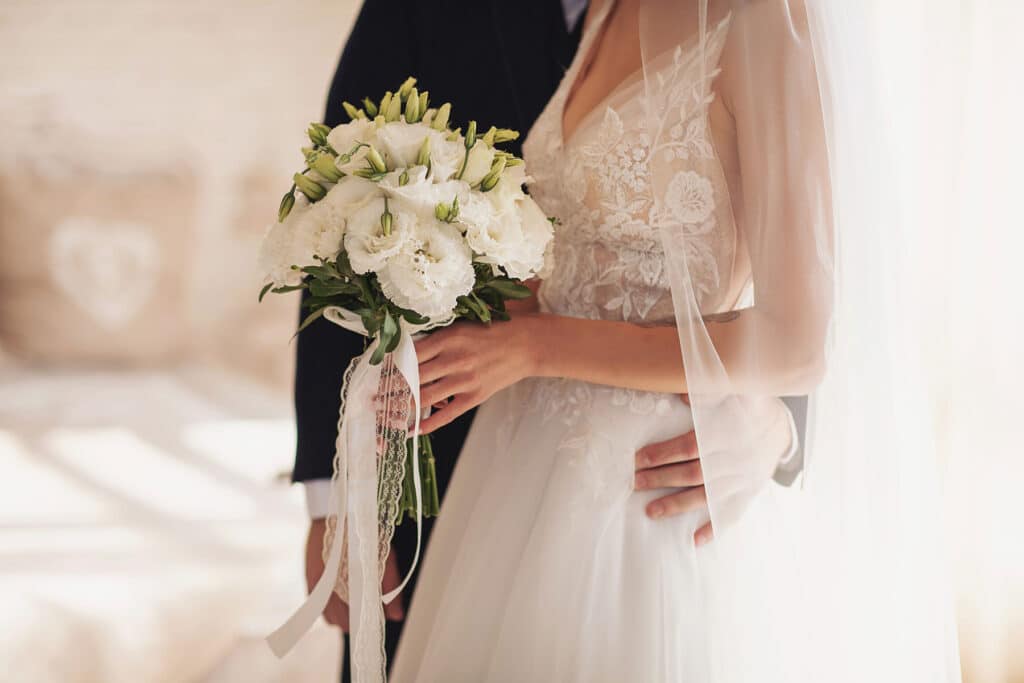 A bride and groom, groom has his hands on her waist and bride is holding a white bouquet.