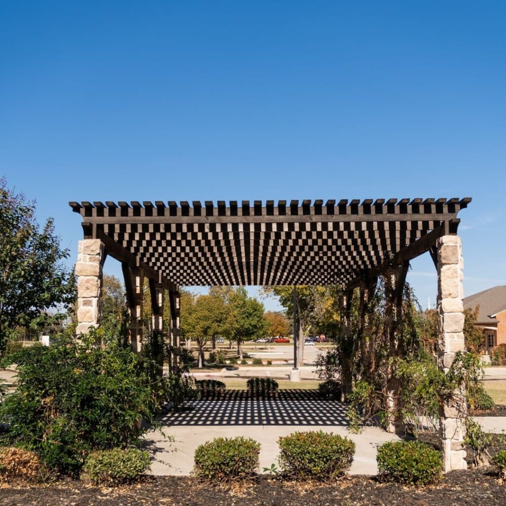 An outdoor wooden pergola with a checkered roof structure and stone columns casting striped shadows on the ground on a sunny day.